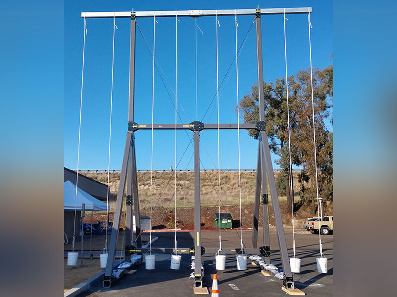 Image shows static load test of the deployable test structure taking place in parking lot. There is a large metal structure raising up and supporting the deployed RF test fixture at two points. The precision test fixture has multiple weights attached across the distance of the structure. 